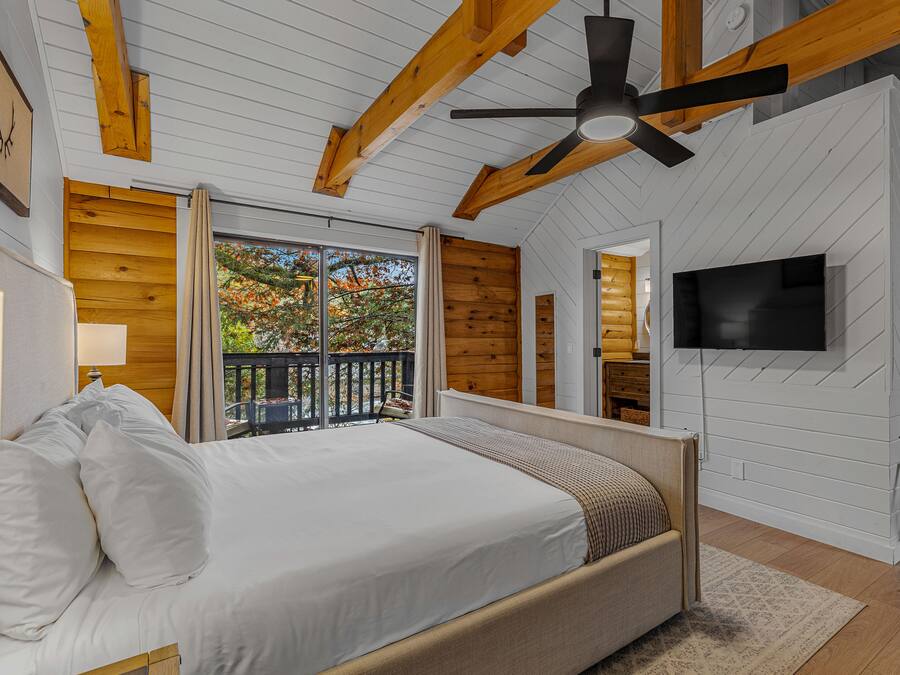 A bedroom with a large bed, wooden ceiling beams, a ceiling fan, a TV mounted on the wall, and a balcony with a view of trees.