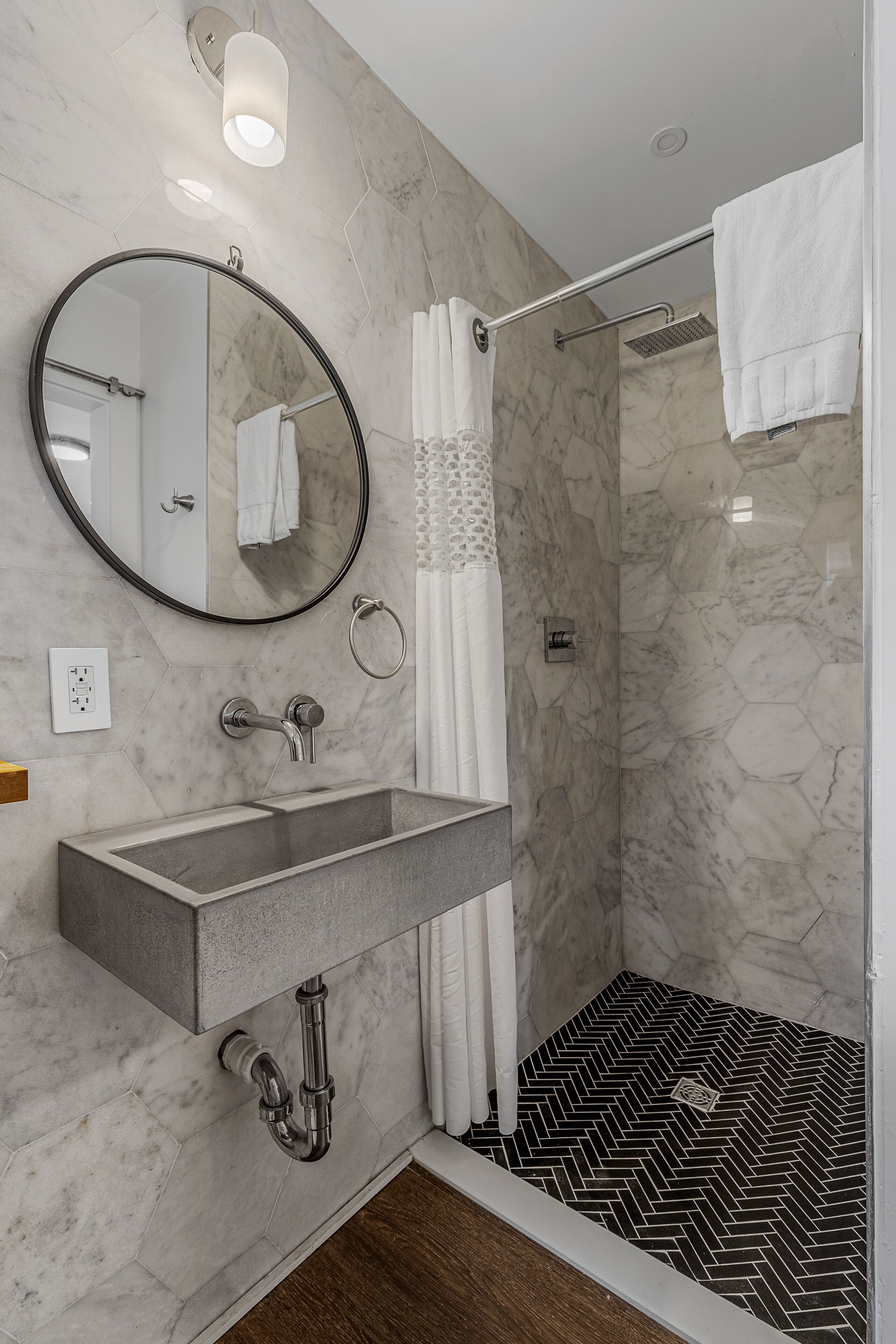 A modern bathroom with a marble wall, a round mirror, a rectangular sink, and a shower with a black and white herringbone floor.