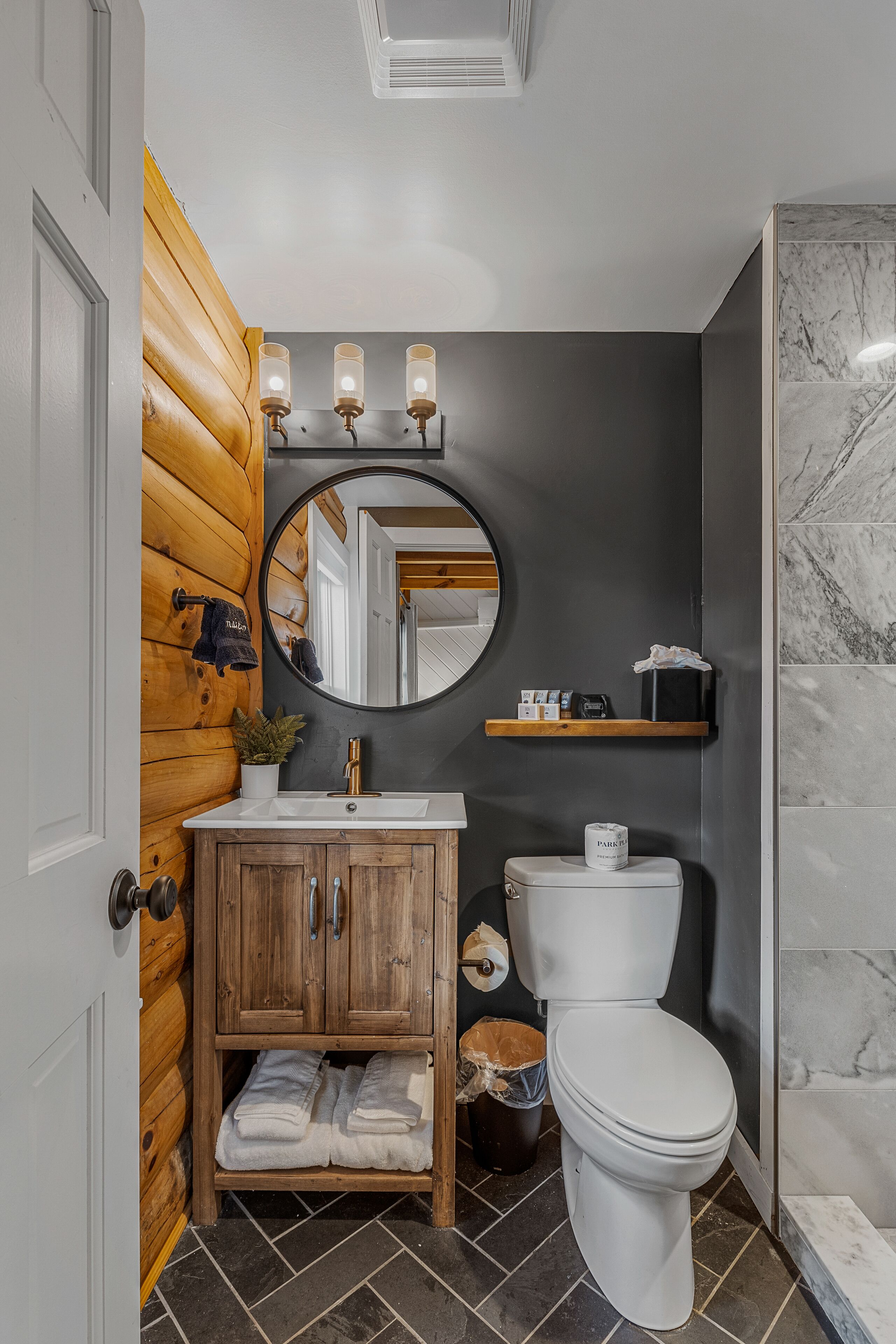 A bathroom with a wooden vanity, a white toilet, a round mirror, and a marble shower area.