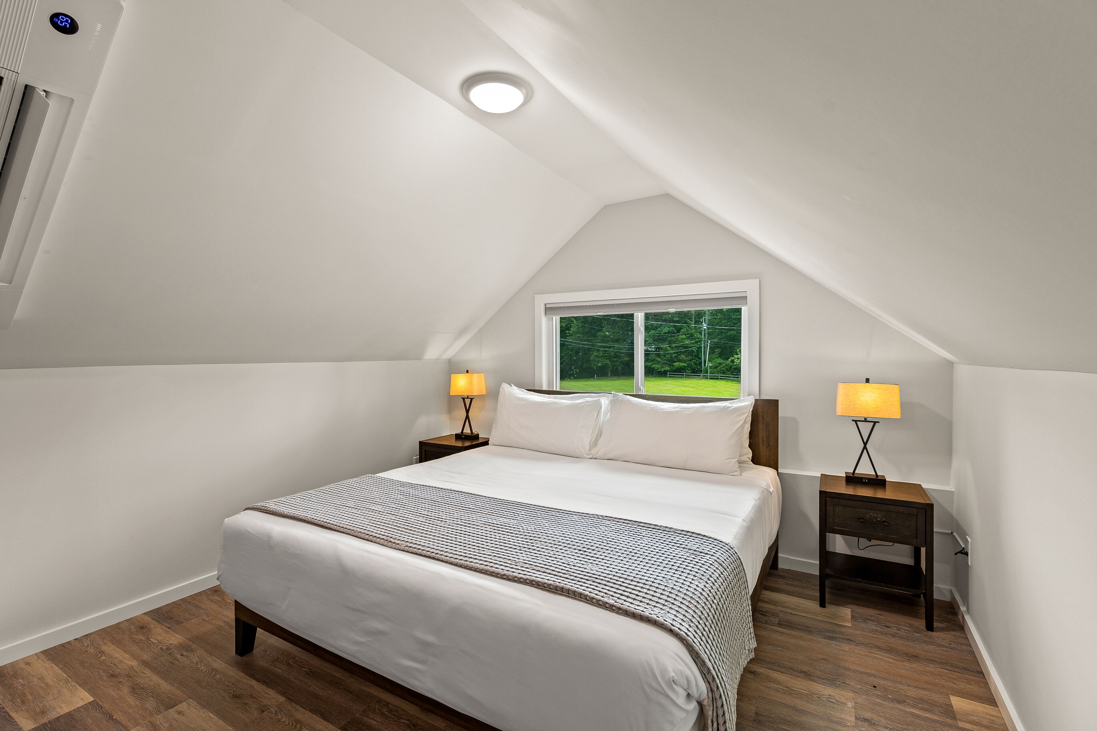 Une chambre avec un plafond en pente, un grand lit, deux tables de chevet avec des lampes et une fenêtre donnant sur un paysage verdoyant.