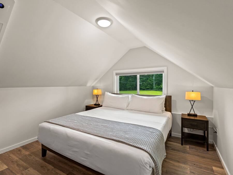 A bedroom with a sloped ceiling, a large bed, two bedside tables with lamps, and a window with a view of greenery.