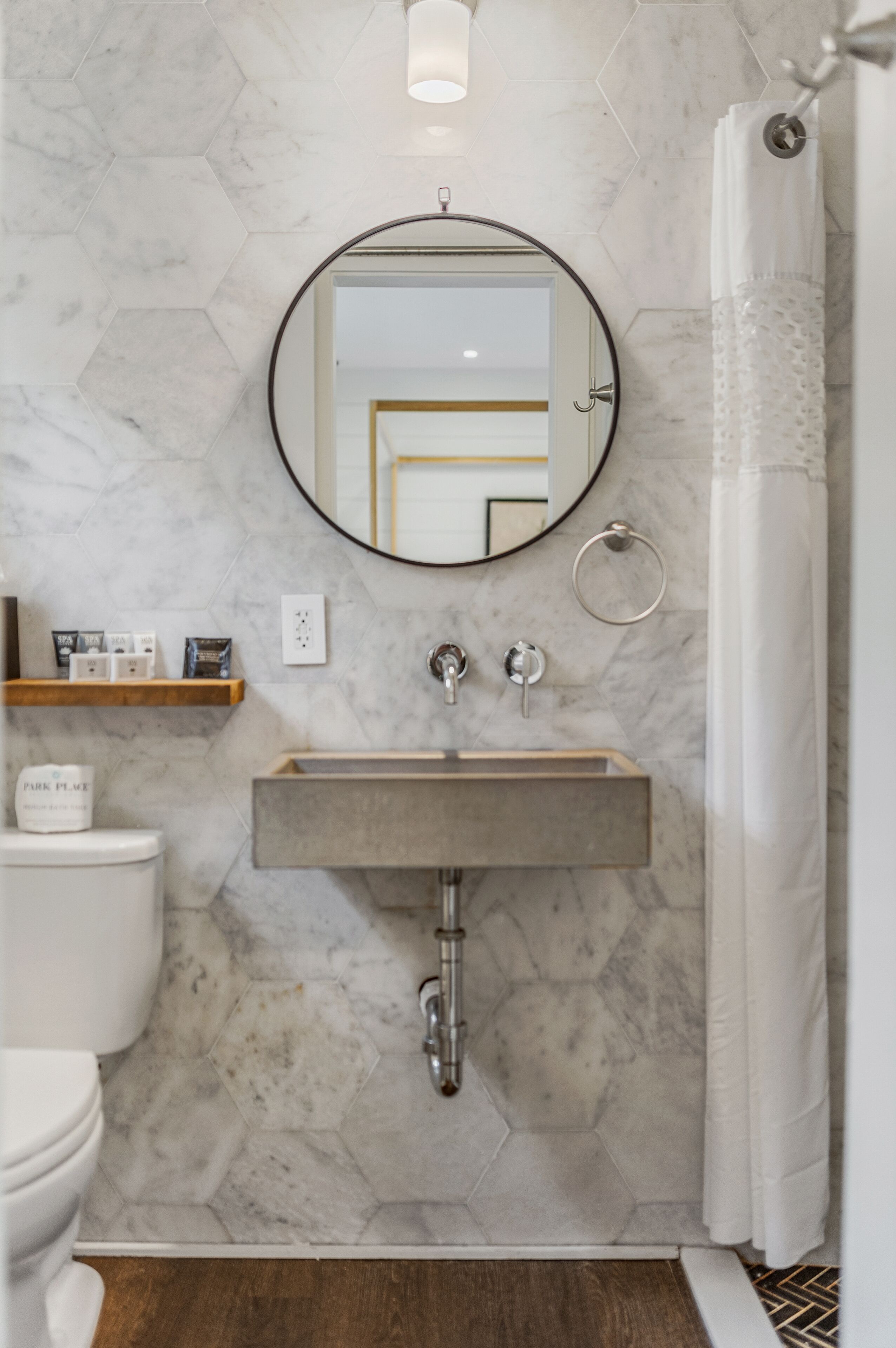 Un baño con un patrón de pared hexagonal de mármol, un espejo redondo, un estante de madera con artículos de tocador, un inodoro y una cortina de ducha.