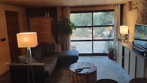 A cozy living room with a sofa, a coffee table, a TV, and a large window with a view of trees.
