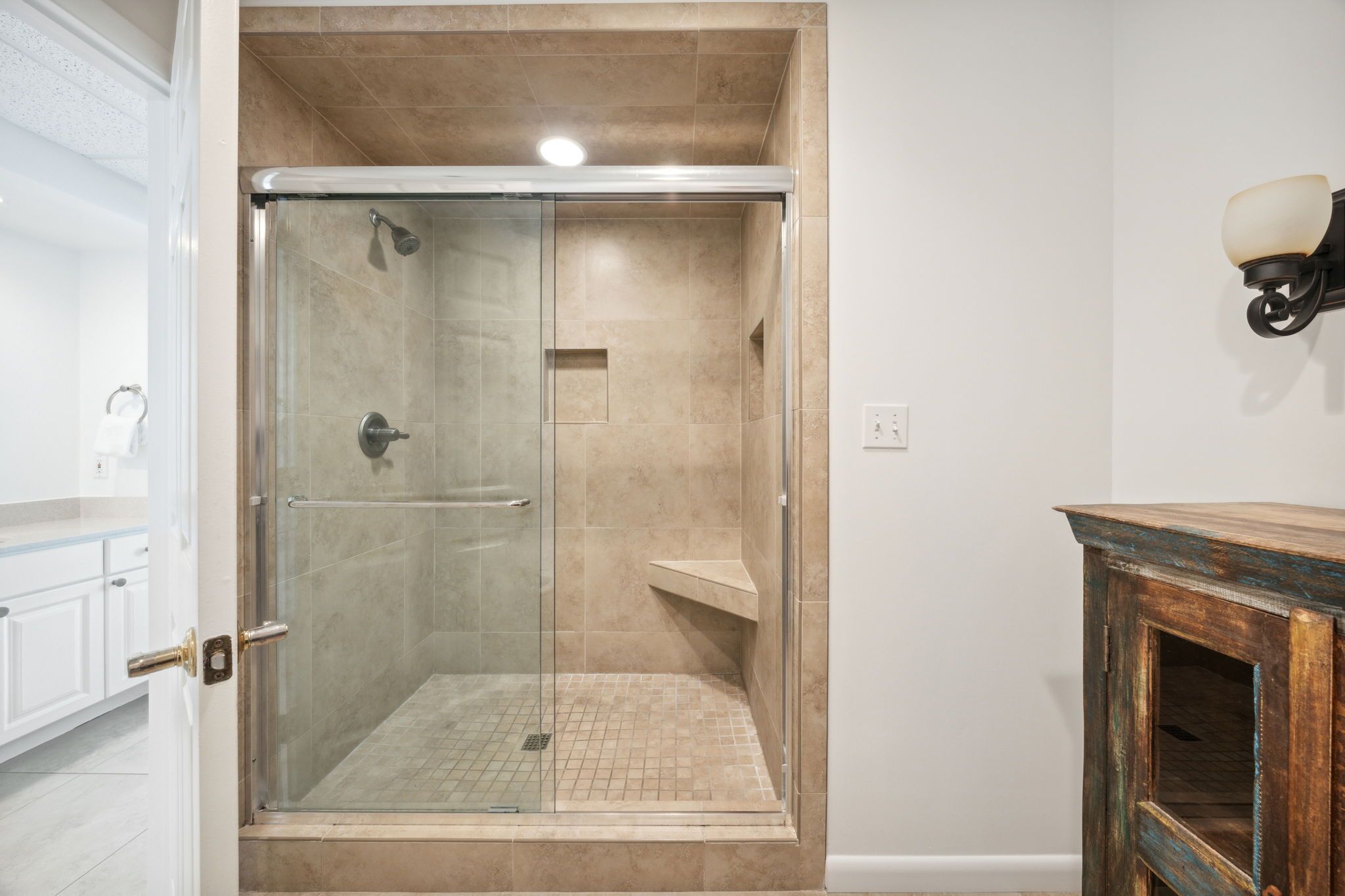 Ducha con cerramiento de vidrio, azulejos color beige, una puerta blanca y un mueble de madera con una lámpara.