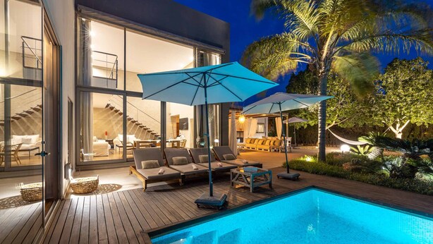 Ein moderner Poolbereich im Freien mit Liegestühlen und Sonnenschirmen, umgeben von einer Holzterrasse und Glasfenstern, die zu einem Innenraum mit einer Treppe führen.