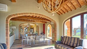 Un vaste espace de vie comprenant une table à manger, des chaises et un canapé. Les murs sont en briques et présentent des arcs, et le plafond est orné de poutres en bois.