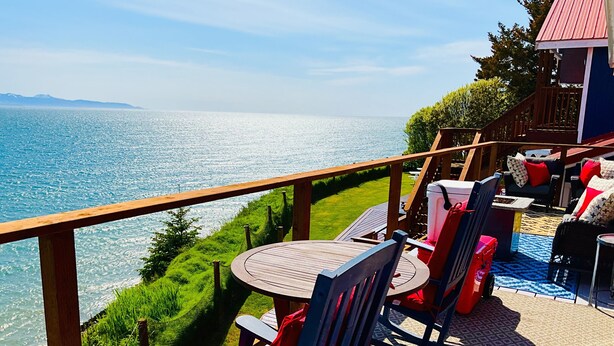 A deck with a view of the sea, featuring a table and chairs, a small building, and a wooden railing. - Oceanfront Two bedroom Condominium  (Homer)