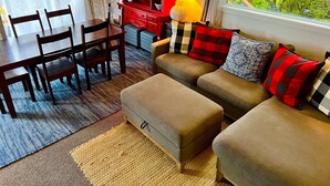 A cozy living room with a sofa, ottoman, and dining table with chairs. A red cabinet with shelves and a lamp is visible. - Oceanfront Two bedroom Condominium  (Homer)