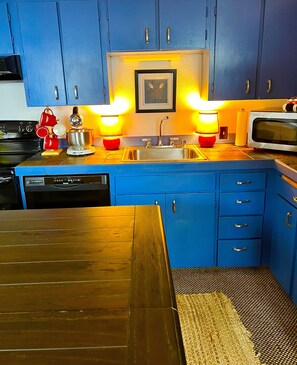 A kitchen with blue cabinets, a stainless steel sink, a microwave, and two red kettles on the countertop.