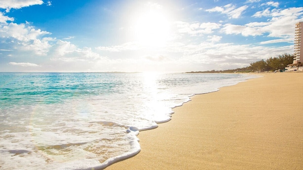 A beach with clear turquoise water, a sandy shore, and a building with multiple balconies in the background.
