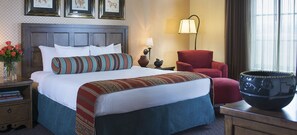 Una cama bien hecha con un almohadón a rayas, un sillón rojo, una lámpara y un florero con flores.