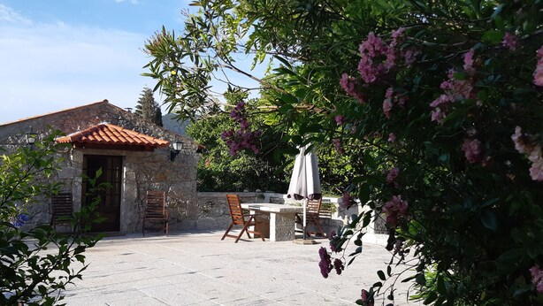En byggnad av sten med ett tak täckt av tegelpannor, ett bord av sten med stolar och en pergola med ett parasoll.
