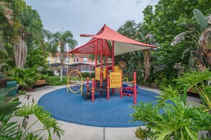 Children can play on the slide and climbing elements in this outdoor play area surrounded by lush greenery and a bench.