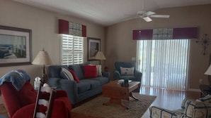 A living room with a blue sofa, red armchair, and a wooden coffee table. There are two lamps, a ceiling fan, and a window with blinds. - Beautiful Villa in the Heart of Florida's Friendliest Active Adult Community (The Villages)
