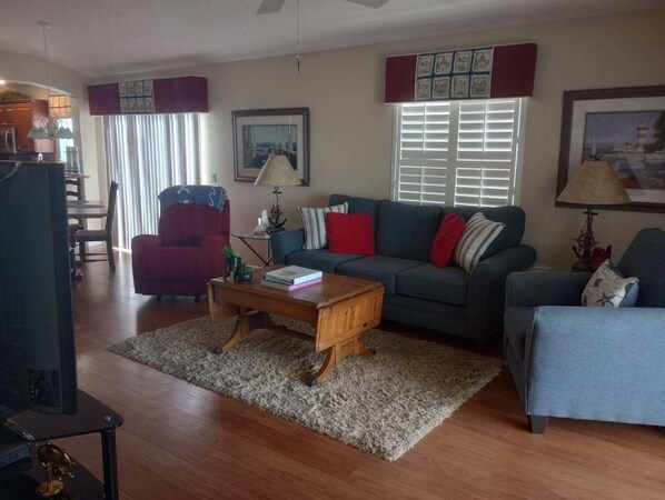A living room with a sofa, armchairs, a coffee table, and a TV. There are framed pictures on the walls and a window with blinds.