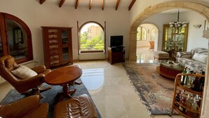 A spacious living room with a high ceiling, wooden beams, a large arched window, a television, leather furniture, and a glass cabinet. - Villa Camelia - Benitachell, Costa Blanca (Benitachell)