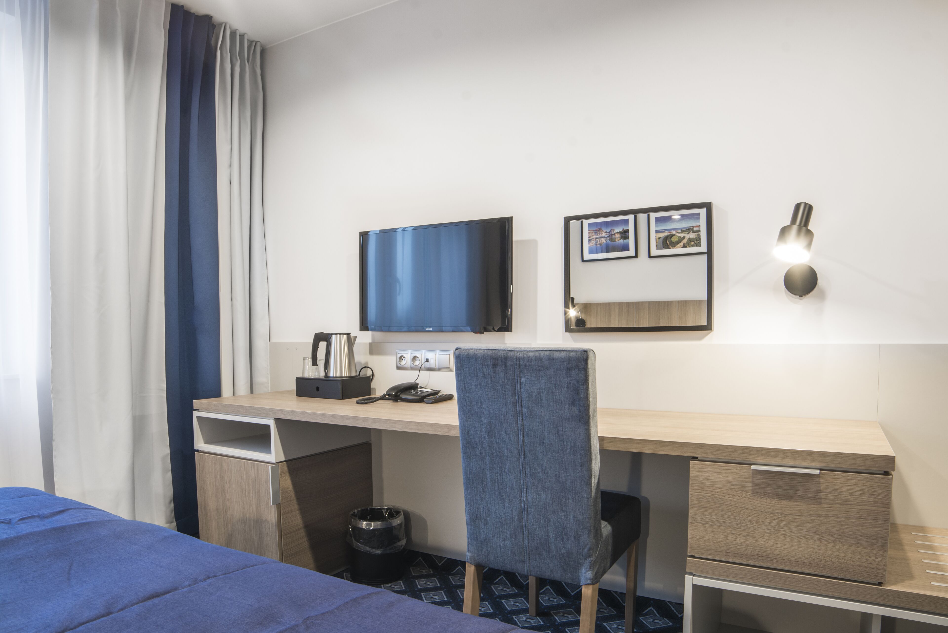Quarto de hotel com cama, escrivaninha, cadeira, televisão, luminária na parede e uma lixeira pequena.