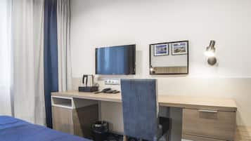 Quarto de hotel com cama, escrivaninha, cadeira, televisão, luminária na parede e uma lixeira pequena.