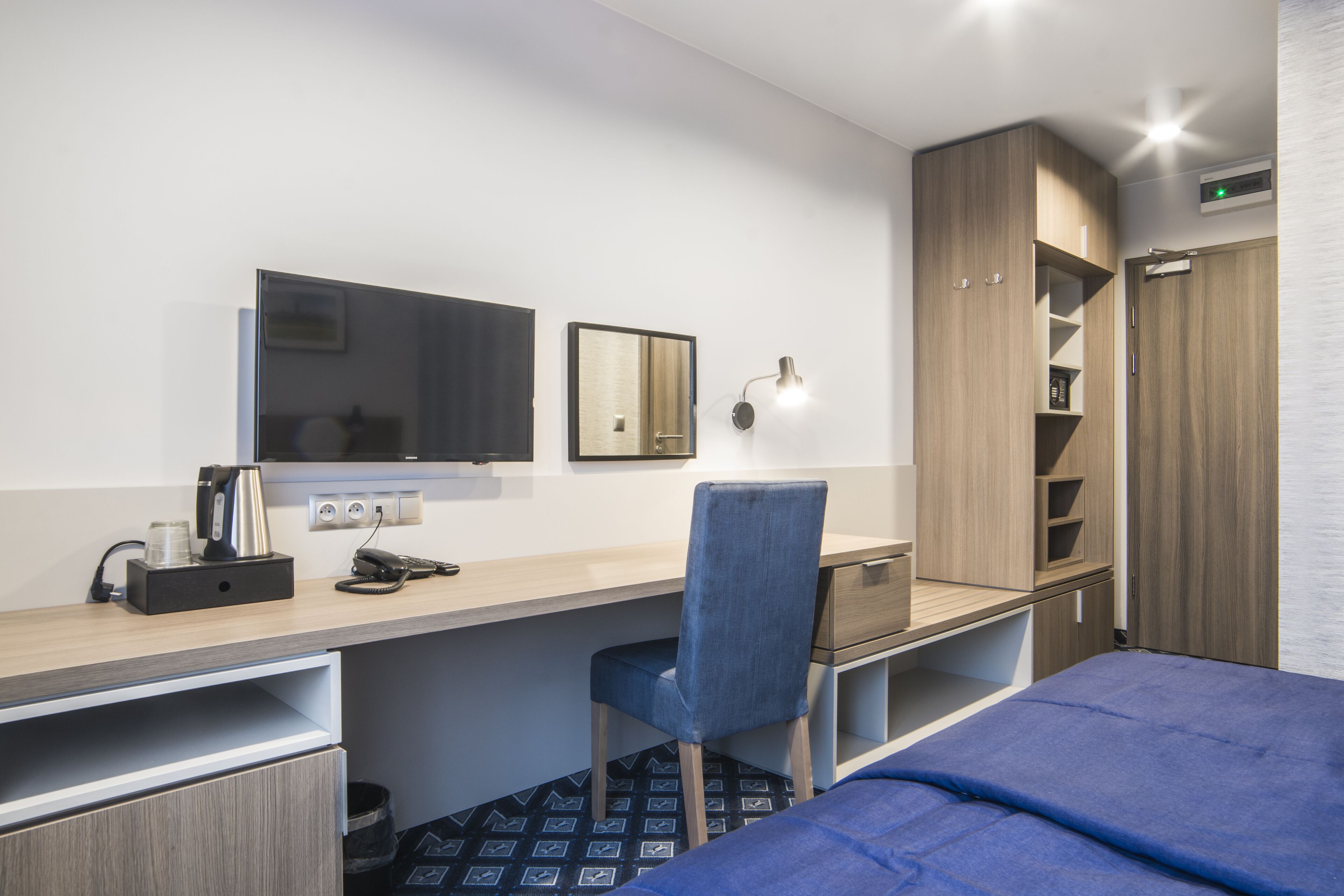 Habitación de hotel con cama, escritorio con silla, espejo, armario y lámpara de pared.