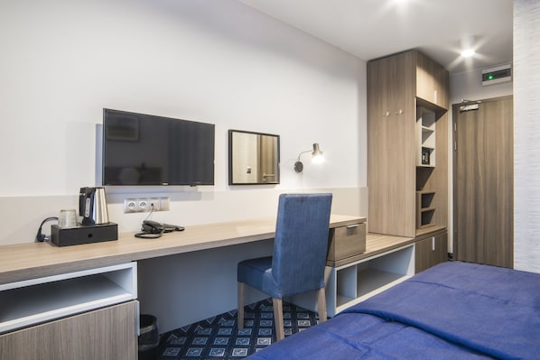 Une chambre d’hôtel comprenant un lit, un bureau avec une chaise, un miroir, une armoire et une lampe fixée au mur.