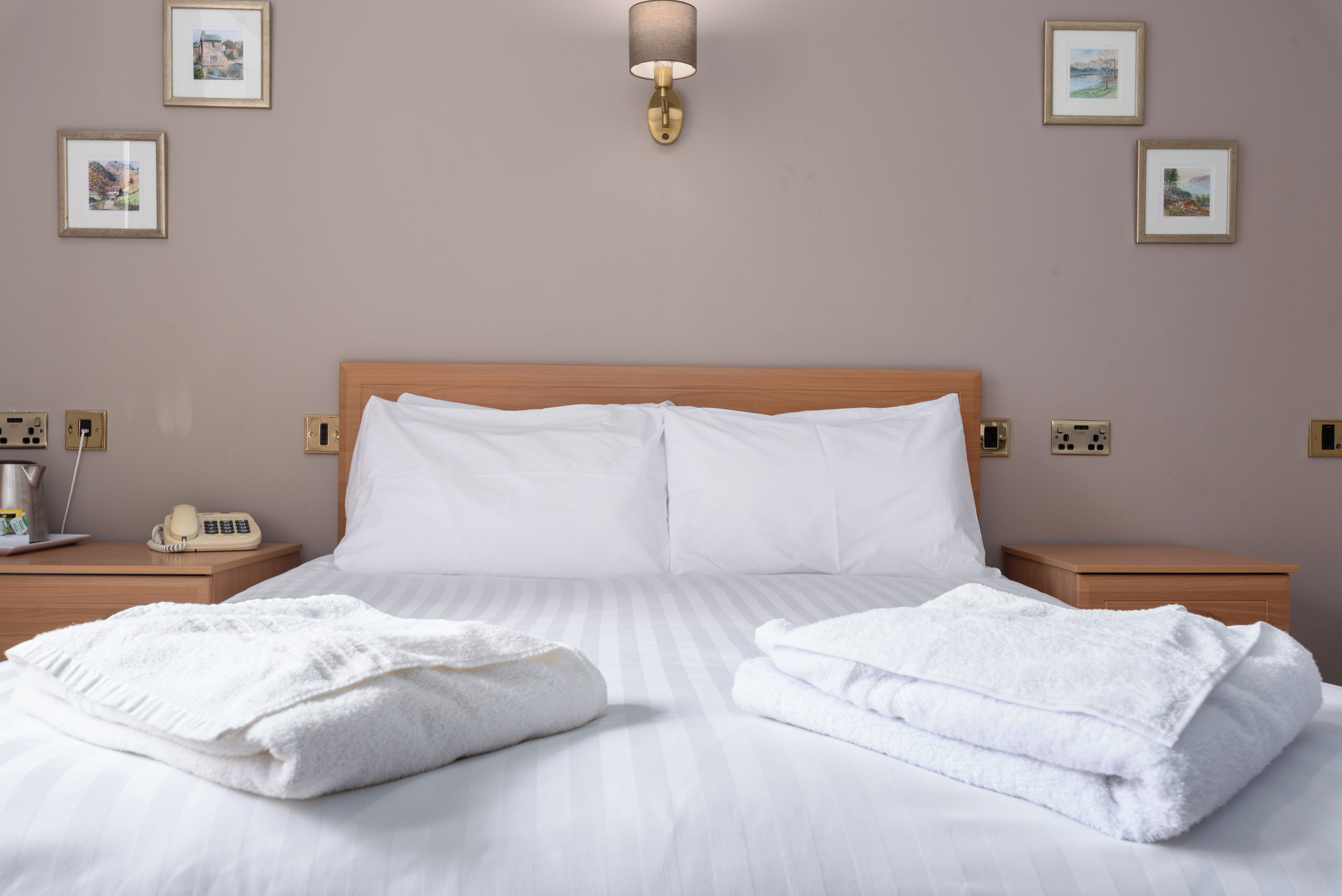 Uma cama bem arrumada com roupa de cama e toalhas brancas, cabeceira de madeira, mesas de cabeceira com uma lâmpada e um telefone, e quadros nas paredes.