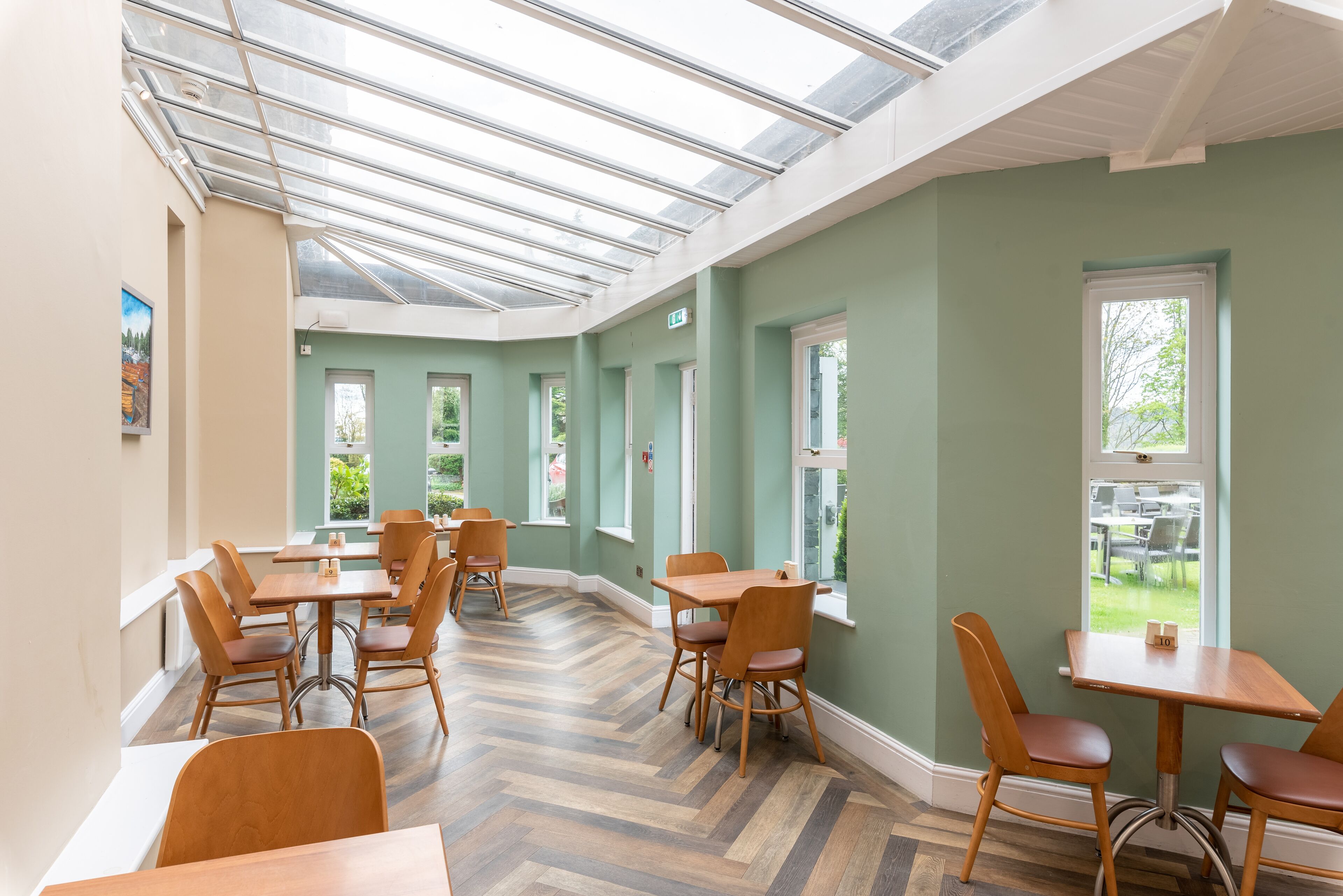 Uma sala de jantar com mesas e cadeiras de madeira, piso com padrão em herringbone e grandes janelas com vista para o verde lá fora.