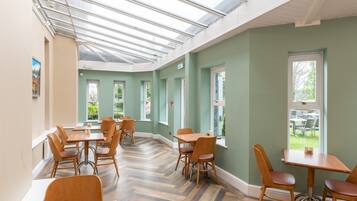 Uma sala de jantar com mesas e cadeiras de madeira, piso com padrão em herringbone e grandes janelas com vista para o verde lá fora.