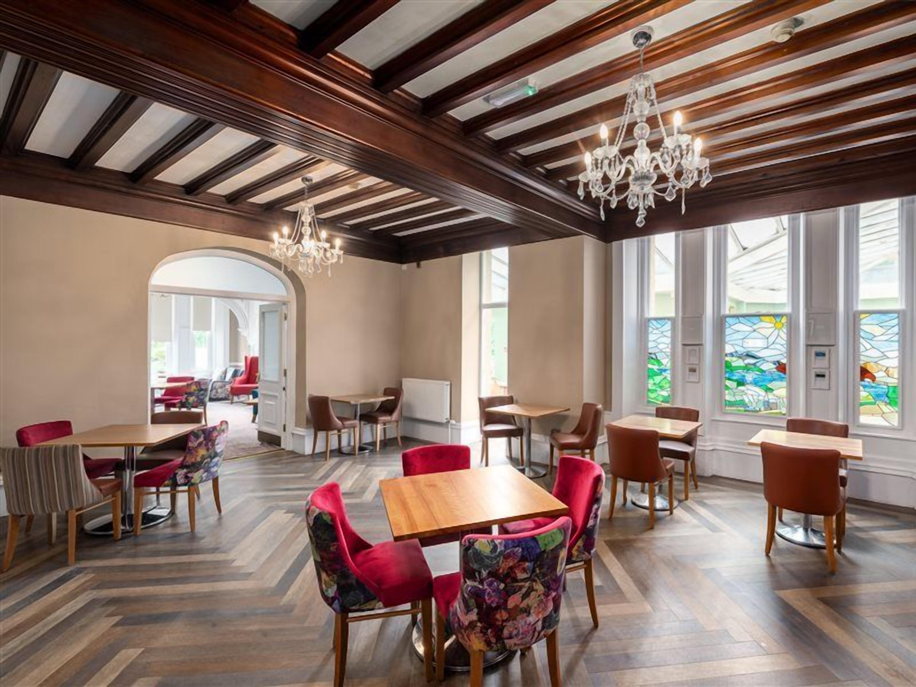 Un comedor amplio con mesas y sillas de madera, un candelabro y ventanas grandes con vidrieras.