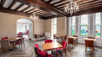 Un comedor amplio con mesas y sillas de madera, un candelabro y ventanas grandes con vidrieras.