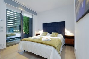 Quarto moderno com uma cama grande, duas mesas de cabeceira, um painel de cabeceira escuro e vista para uma parede verde através de uma porta deslizante.