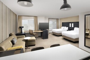 Quarto de hotel com sofá, duas camas de solteiro, uma mesa de trabalho com cadeira, televisão e uma janela grande.