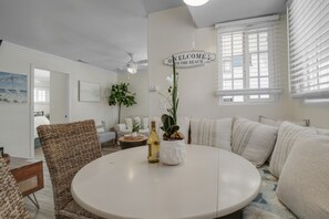A living room with a round table, wicker chair, and a sofa. There is a 'WELCOME TO THE BEACH' sign on the wall.