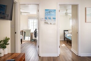 A hallway leading to a bedroom with a bed, a desk with a chair, and a painting of a beach scene.