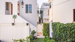 Une ruelle étroite bordée d’une clôture blanche, un petit jardin avec différentes plantes et un bâtiment blanc doté d’un toit en tuiles.