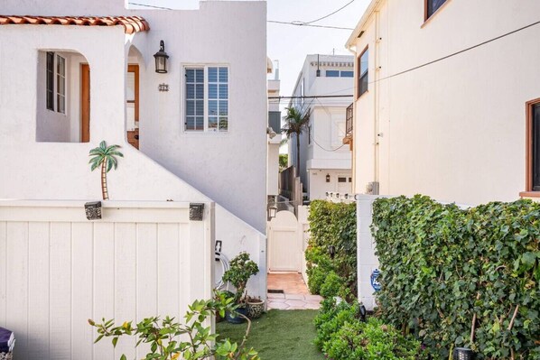 Uma viela estreita com cerca branca, um pequeno jardim com diversas plantas e um edifício branco com telhado em tijolos.