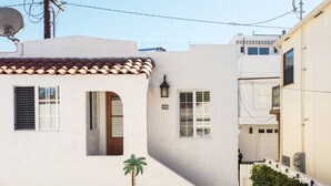 Un bâtiment blanc avec un toit en tuiles, un petit balcon orné de plantes et un décor composé de palmiers.
