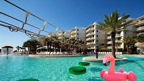 Una piscina con sillones de descanso, palmeras y un edificio con balcones al fondo.