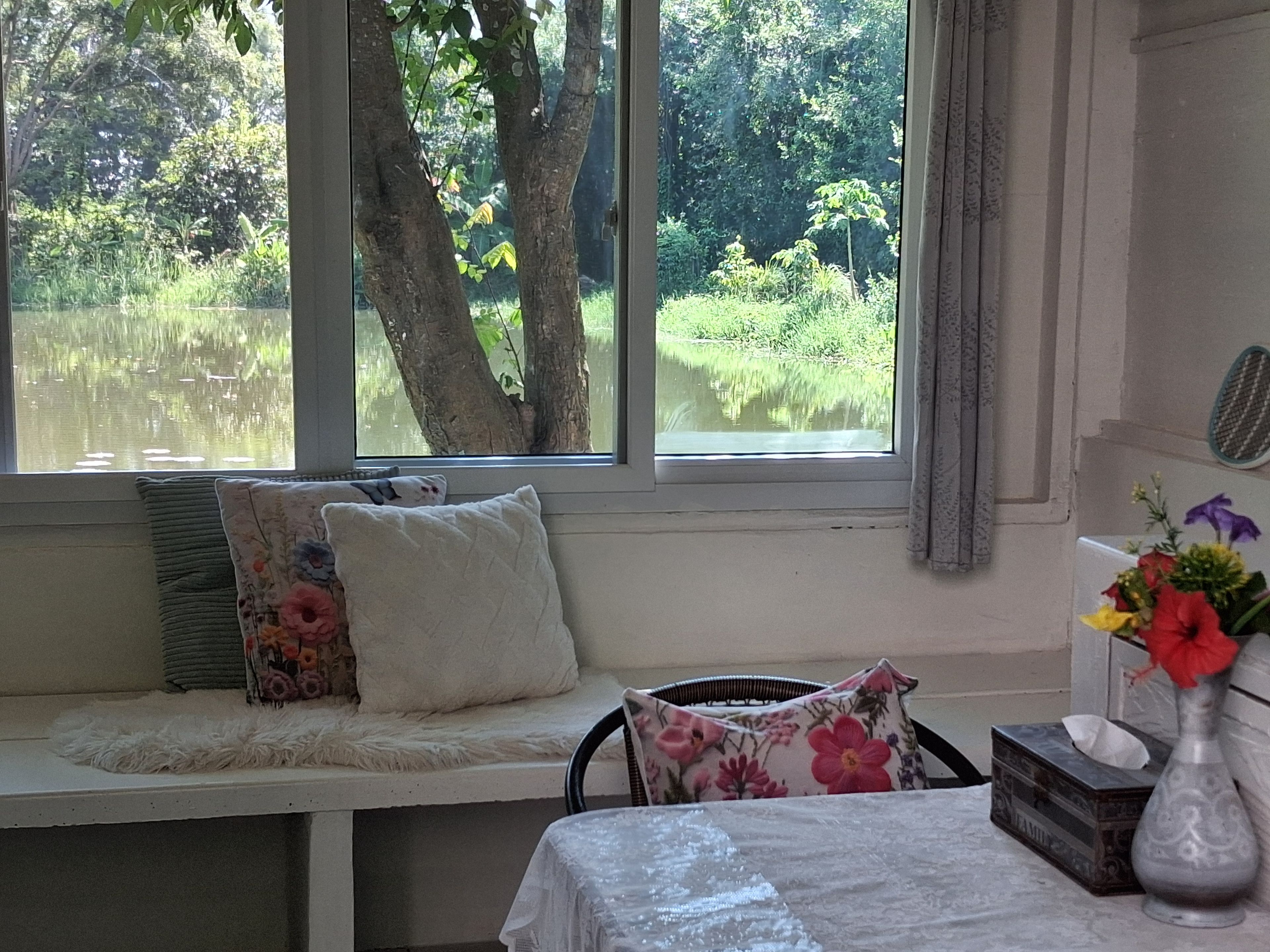 A room with a window seat overlooking a pond, a table with a vase of flowers, and a tissue box.