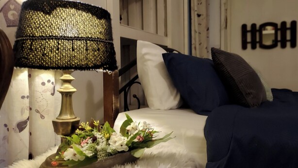 A neatly made bed with white and dark blue bedding, a decorative lamp, and a floral arrangement on a side table. - Village Homestay (Kumphawapi)