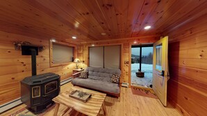 A cozy cabin interior with a wood stove, sofa, and a view of a snowy landscape through the door.