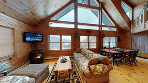 A log cabin interior with a large fireplace, a sofa, a dining table, and a TV mounted on the wall.