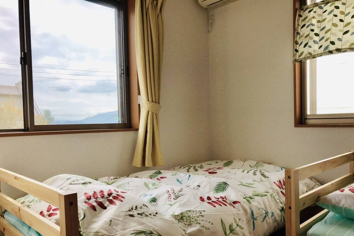 Une petite chambre avec un lit simple, un cadre en bois, une couvre-lit à motifs floraux et une vue sur l’extérieur par une fenêtre ornée d’un rideau.