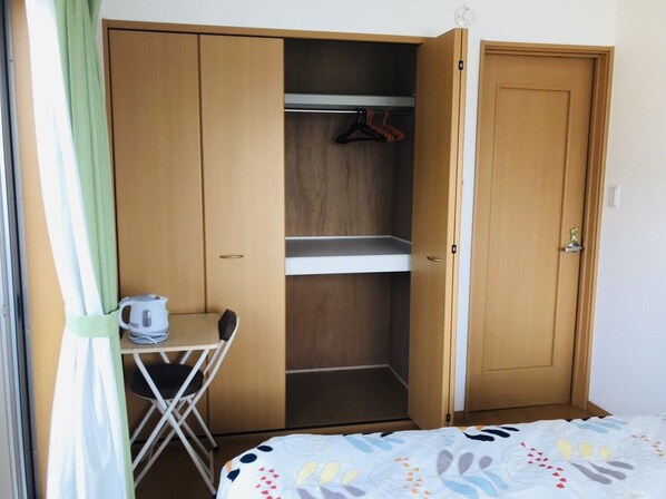 Une chambre avec un lit, un petit bureau avec une bouilloire, une chaise et une armoire avec une étagère.