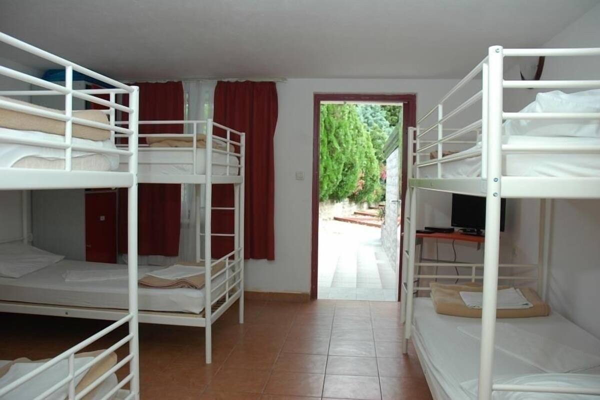 A dormitory room with bunk beds, a television on a desk, and a view of a garden through an open door.