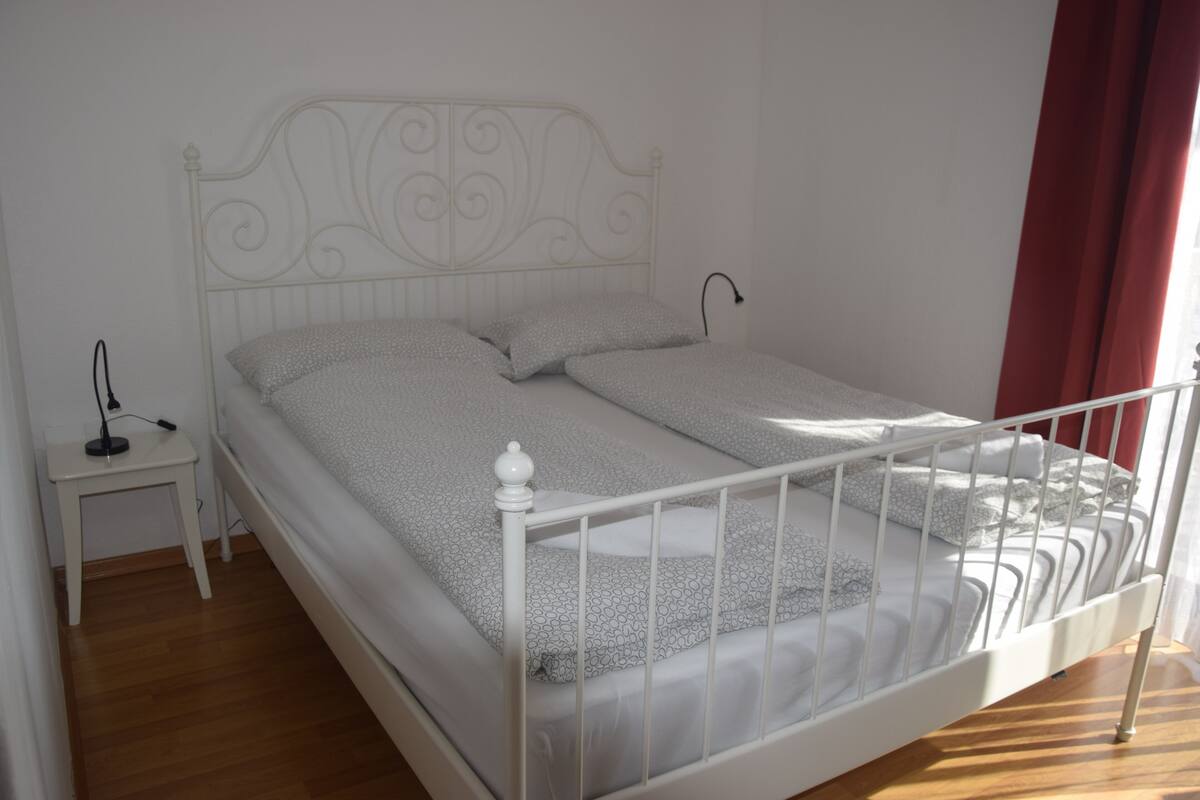 A white metal bed with a decorative headboard, a small bedside table, and a red curtain.