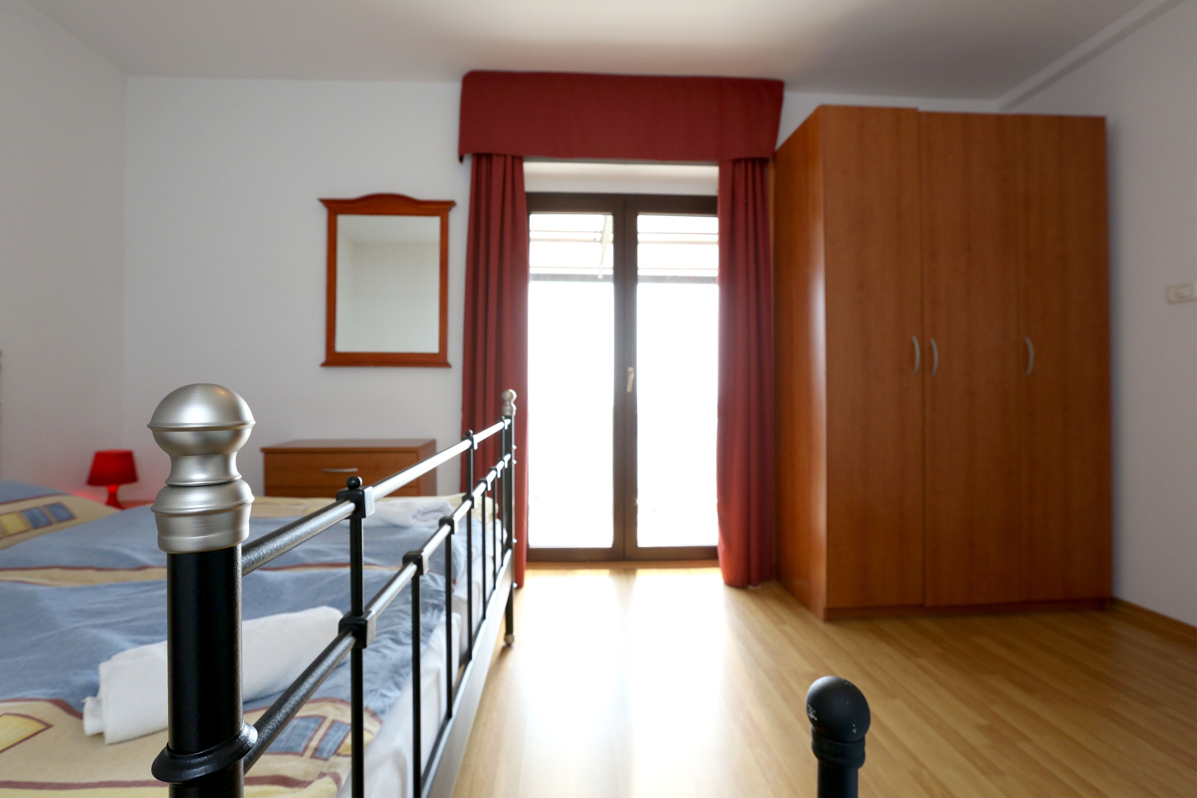 A bedroom with a bed, a wooden wardrobe, a mirror, and a door leading outside.