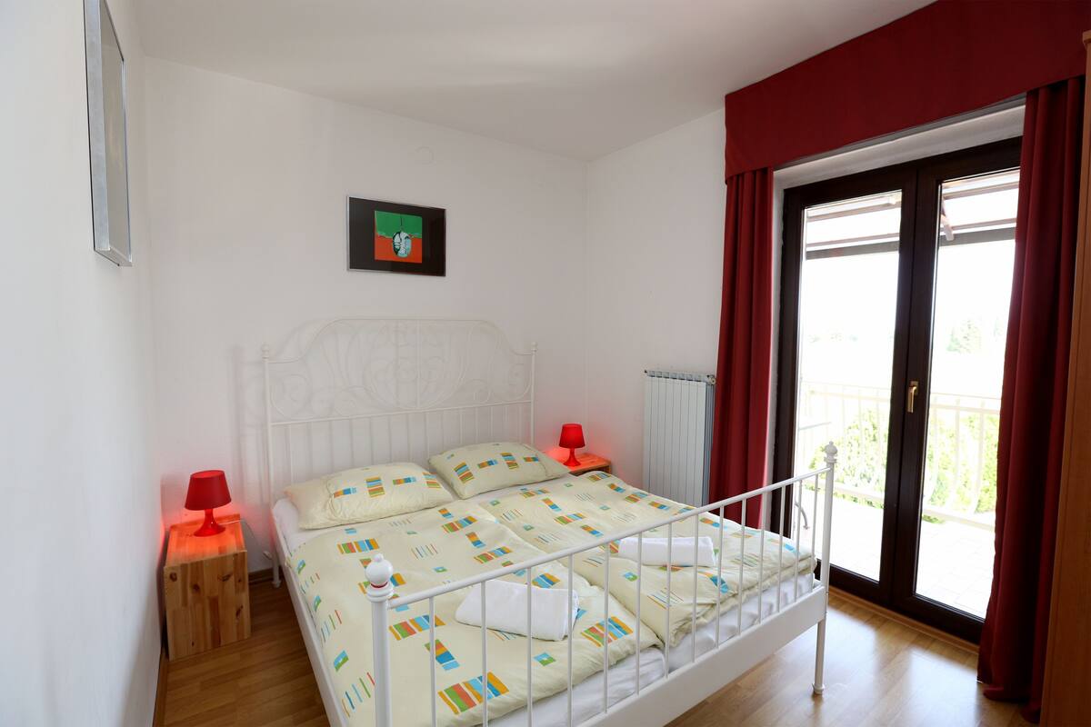 A bedroom with a white bed, red curtains, a small bedside table with a lamp, and a window with a view of greenery.