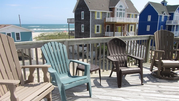 Une terrasse en bois, agrémentée de chaises Adirondack et d’un banc, offre une vue sur une plage, avec des maisons en arrière-plan.