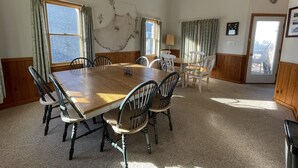 Une salle à manger avec une grande table et des chaises en bois, un ventilateur de plafond et une vue sur l’extérieur par les fenêtres.
