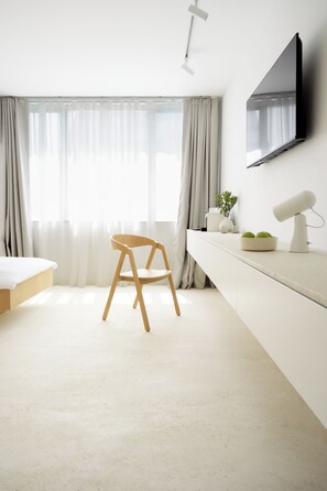 Habitación de hotel minimalista con cama, escritorio con lámpara, una silla y televisor montado en la pared.